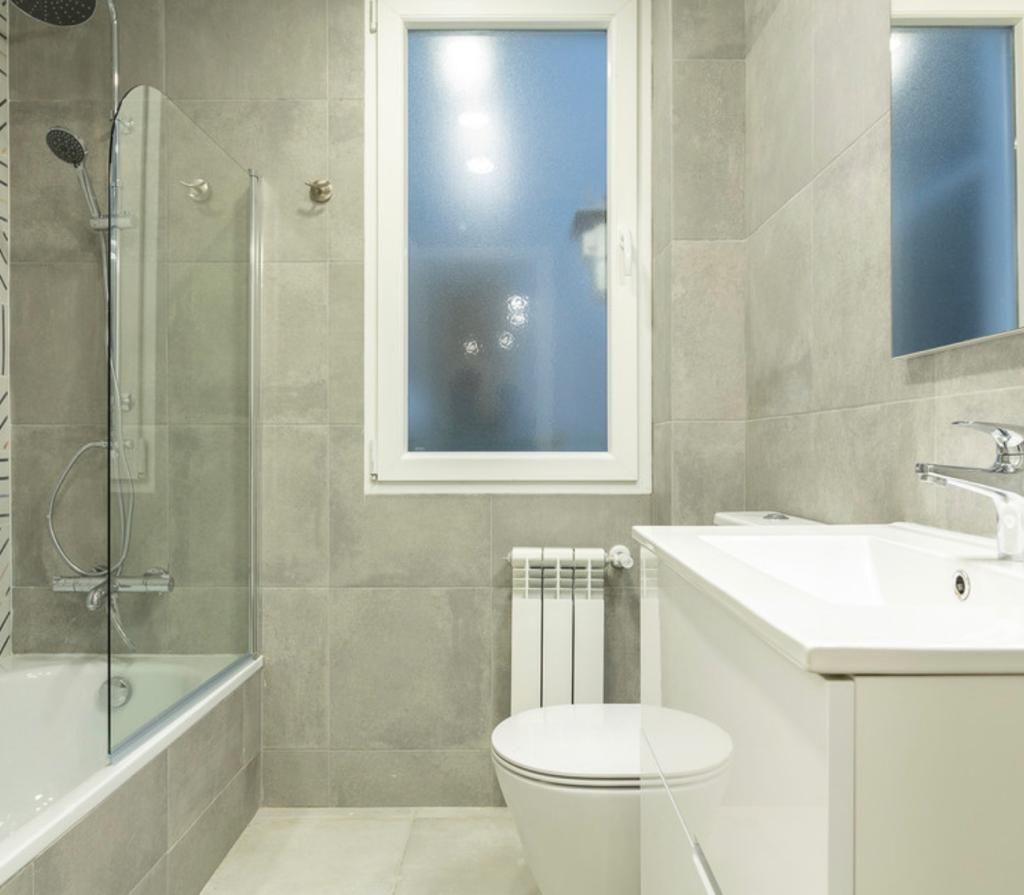 Baño reformado con revestimiento cerámico gris en las paredes, ducha con mampara y lavatorio blanco en un departamento de CABA